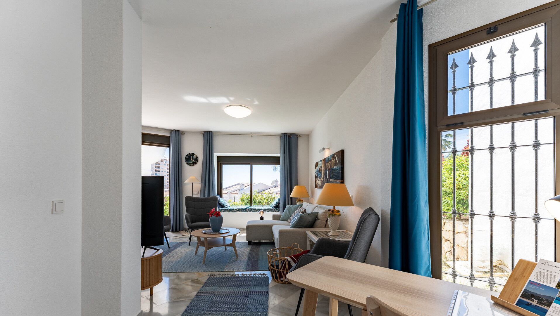 A spacious living room with a sofa, armchairs, a coffee table, and a TV. There is a large window with curtains and a view of the outdoors.
