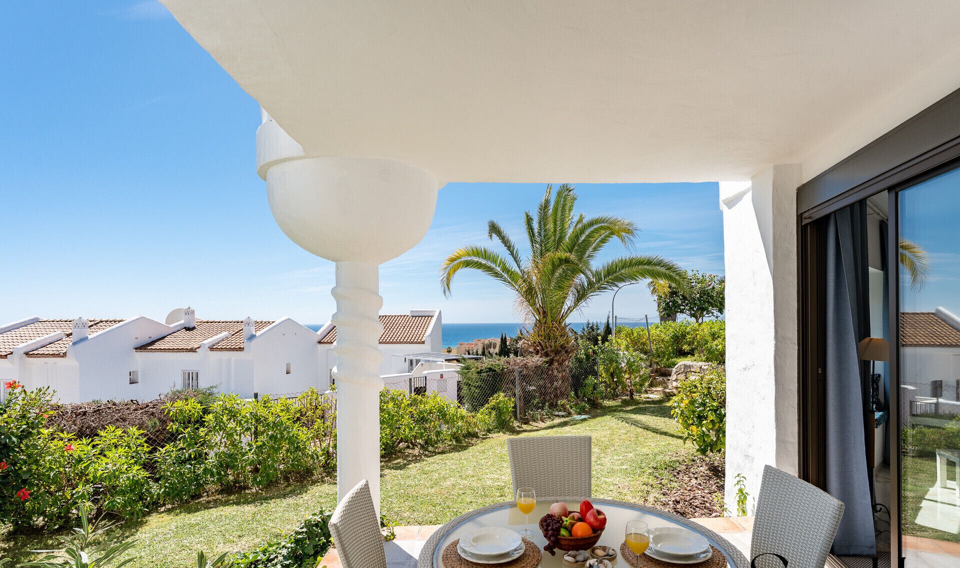 A patio with a table set for two, overlooking a garden with a palm tree and a view of the sea.