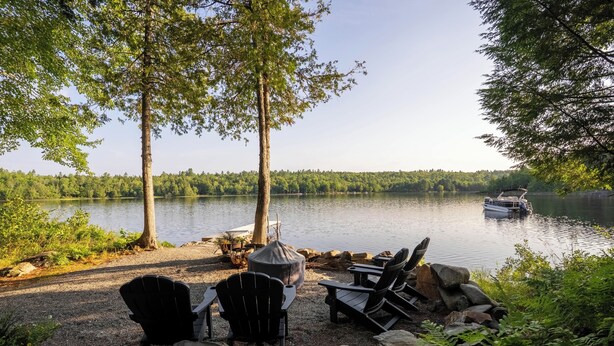 Un área de estar junto al lago con sillas Adirondack, una parrilla cubierta y un bote en el agua.
