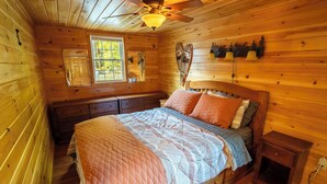 Dormitorio de cabaña de madera con cama, mesita de noche, ventana y ventilador de techo.