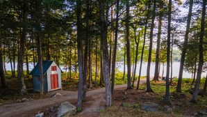 Una cabaña con puerta roja y techo azul, rodeada de árboles altos y un sendero de tierra que lleva a un lago.