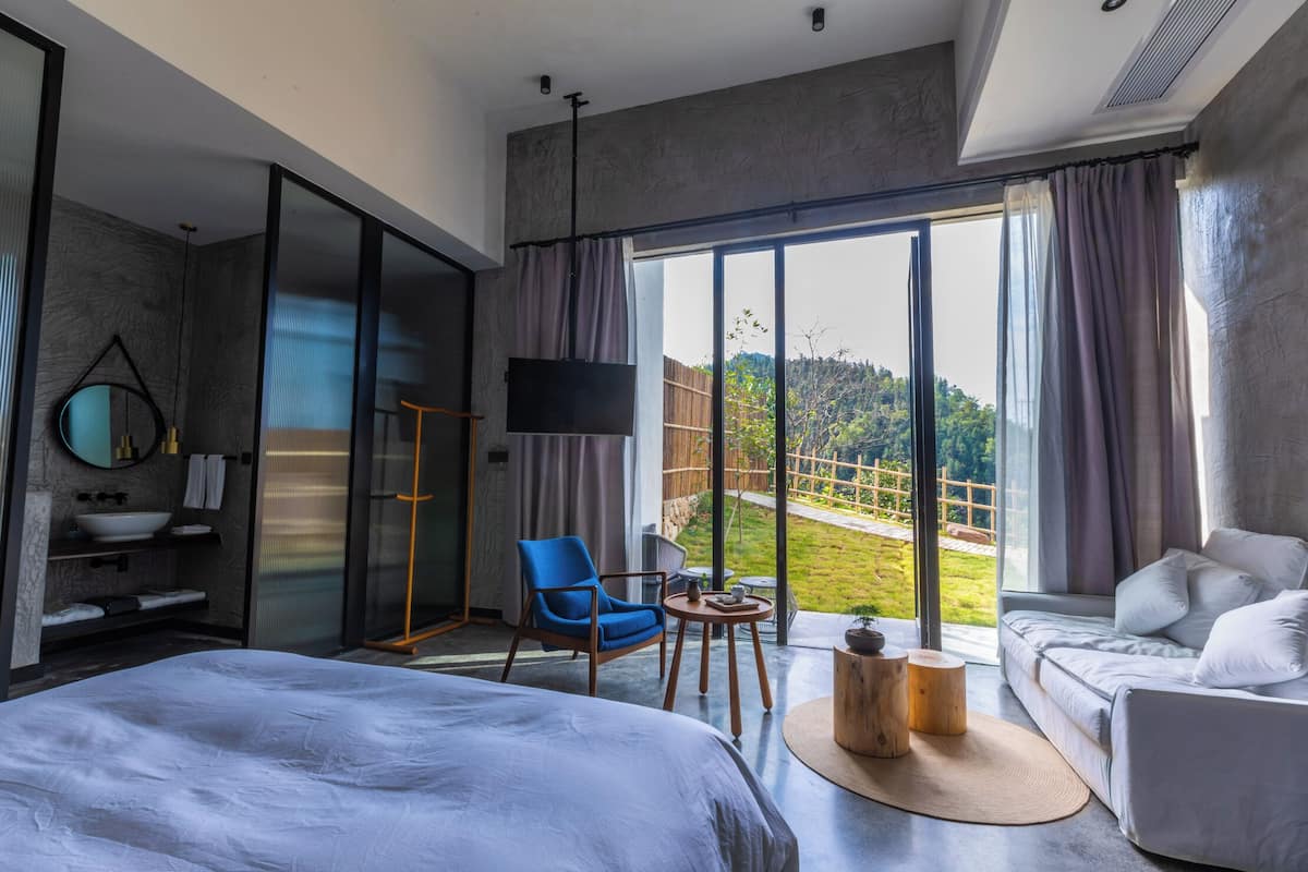 Ett modernt hotellrum med en stor säng, en sittgrupp med soffa och fåtölj, ett litet bord och ett stort fönster med utsikt över omgivningarna.