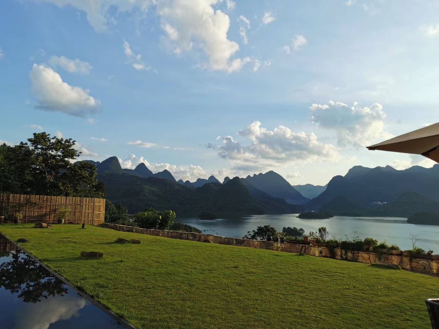 Zona verde con un muro de piedra, una cerca de madera y vistas a montañas y un lago.