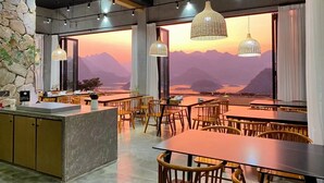 Un restaurante con vistas a las montañas y al atardecer. Tiene mesas y sillas de madera, y luces colgantes.