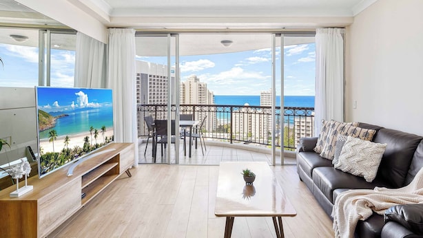 A modern living room with a large TV, a sofa, a coffee table, and a balcony with a view of the sea and buildings. - Chevron Renaissance Ocean View (Surfers Paradise)