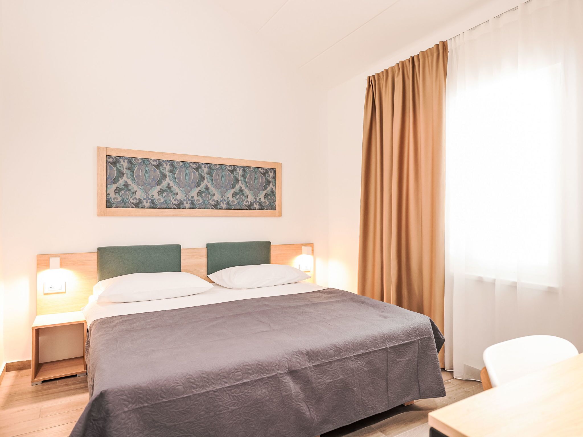 Een keurig opgemaakte hotelkamer met een bed, twee nachtkastjes met lampen, een lijst met kunstwerk aan de muur en een raam met gordijnen.