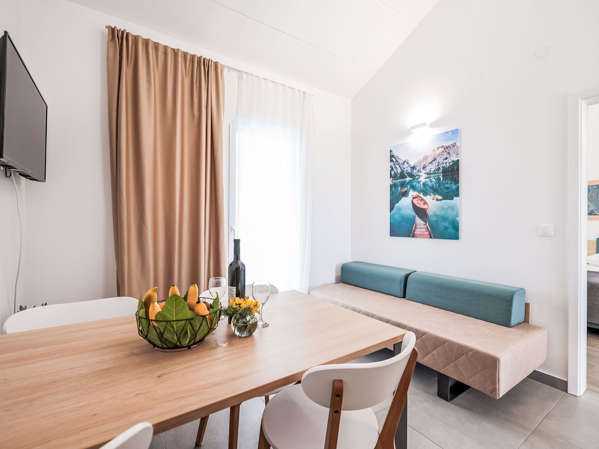 A modern room with a wooden dining table, a bench, a wall-mounted TV, and a painting of a mountain landscape.