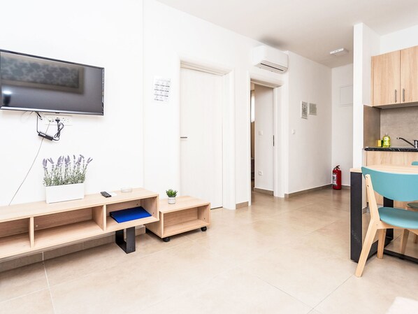 A modern, well-lit room with a flat-screen TV mounted on the wall, a small plant on a shelf, a desk with a chair, and a kitchenette area with cabinets and a sink.