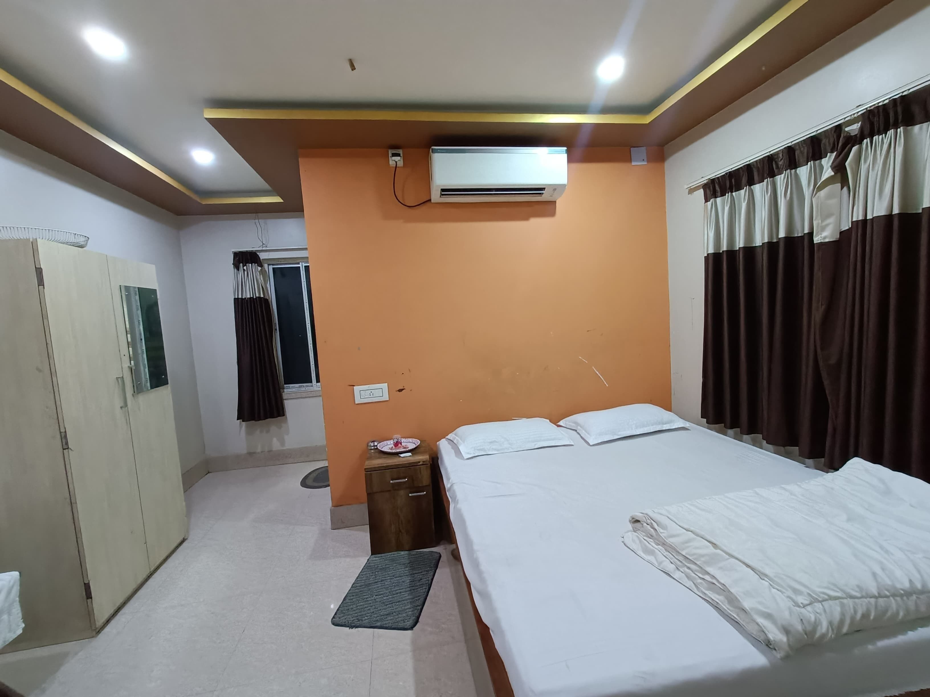 Habitación de hotel con cama, armario, mesita pequeña, equipo de aire acondicionado y ventana con cortinas.