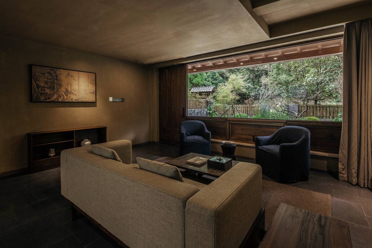 A modern living room with a sofa, two armchairs, a coffee table, and a large window offering a view of a garden.