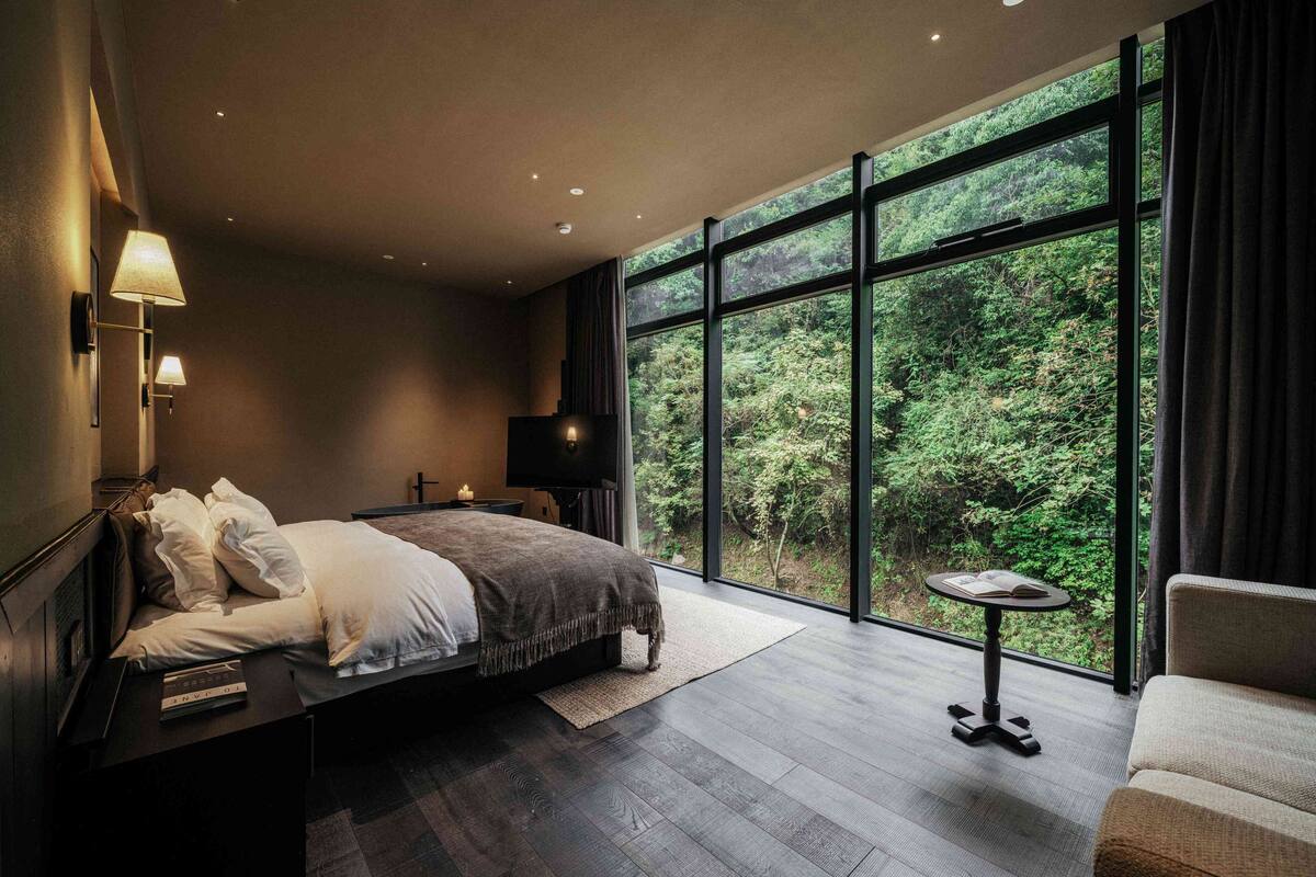 A modern bedroom with a large window, a bed, a bedside table, a TV, and a small table with a chair.