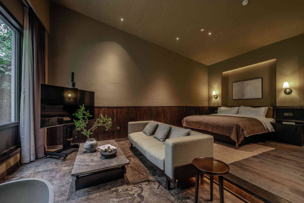 A modern hotel room with a large bed, a sofa, a small table, a TV, and a bathroom.