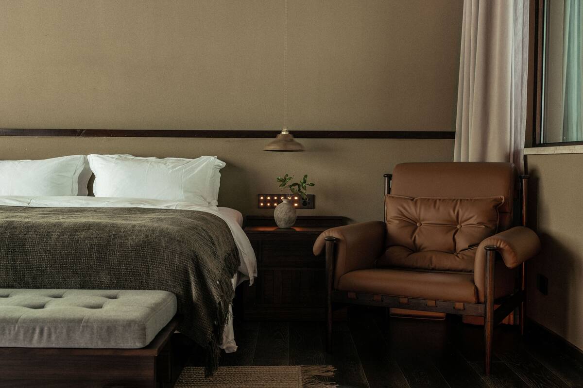 A bedroom with a bed, a nightstand with a lamp, a chair, and a window with curtains.