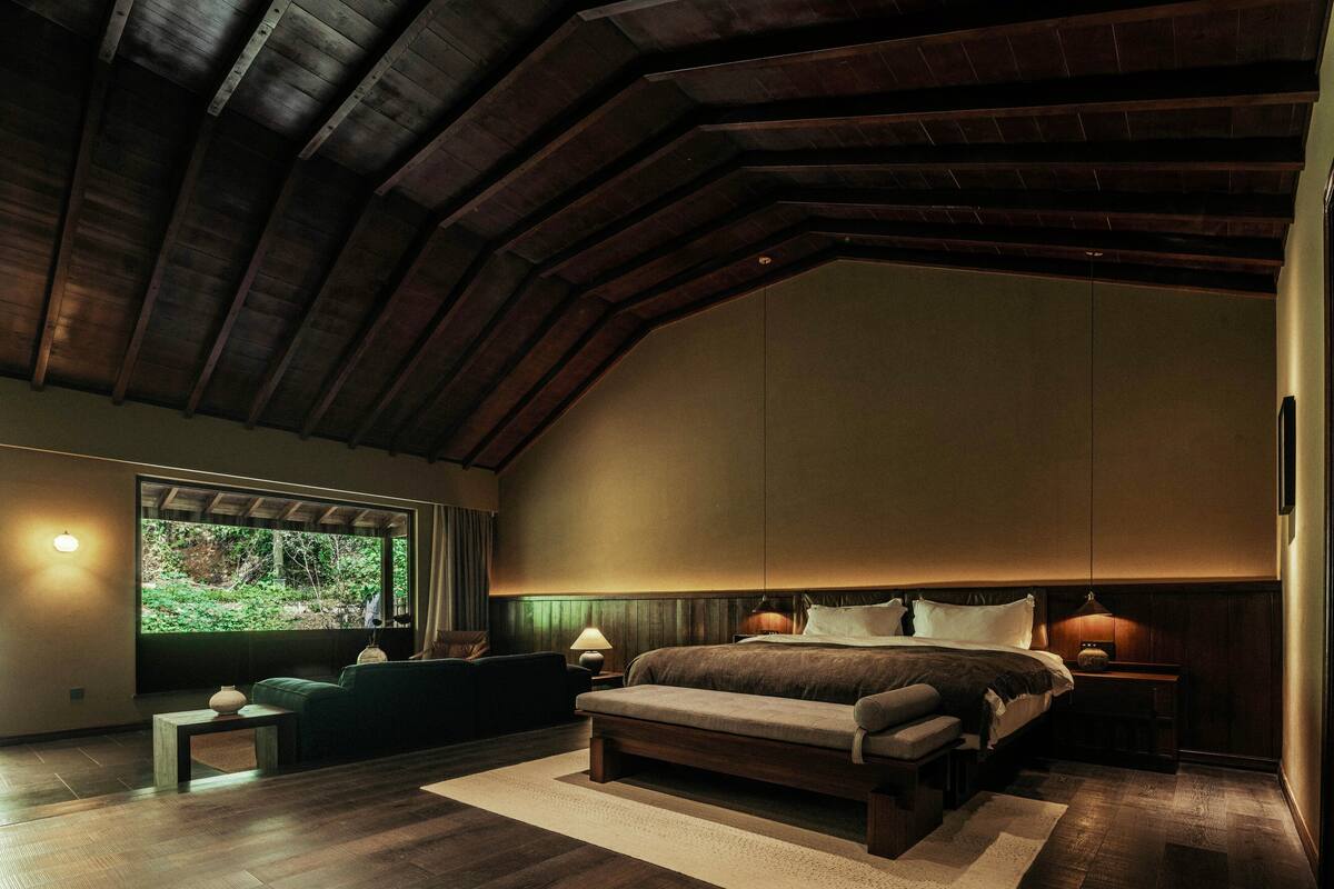A bedroom with a large bed, a bench, a nightstand, and a window with a view of greenery.