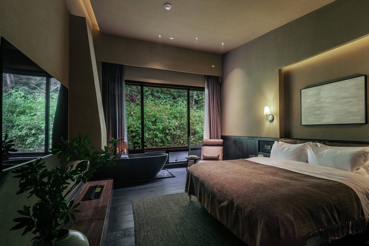 A hotel room with a large bed, a sitting area with a table and chair, a large window with a view of greenery, and a wall-mounted lamp.