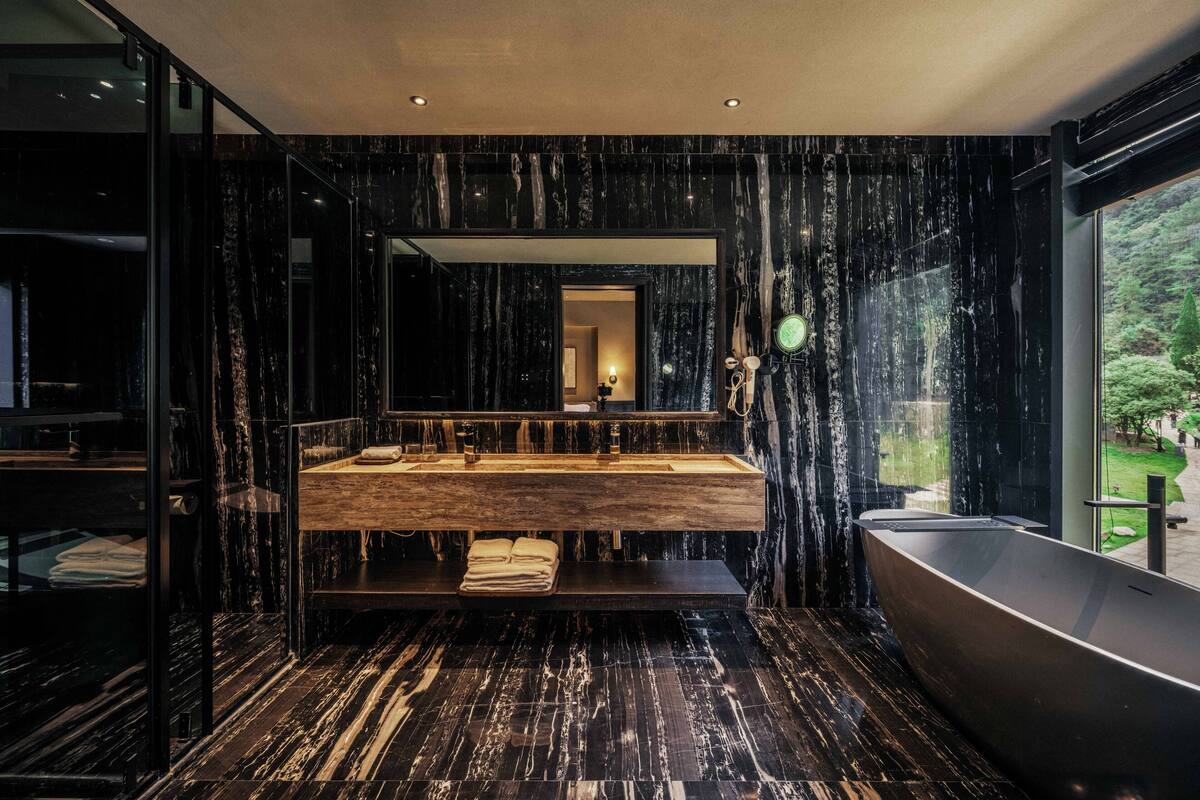 A modern bathroom with a large mirror, a wooden vanity, a bathtub, and a view of greenery outside.