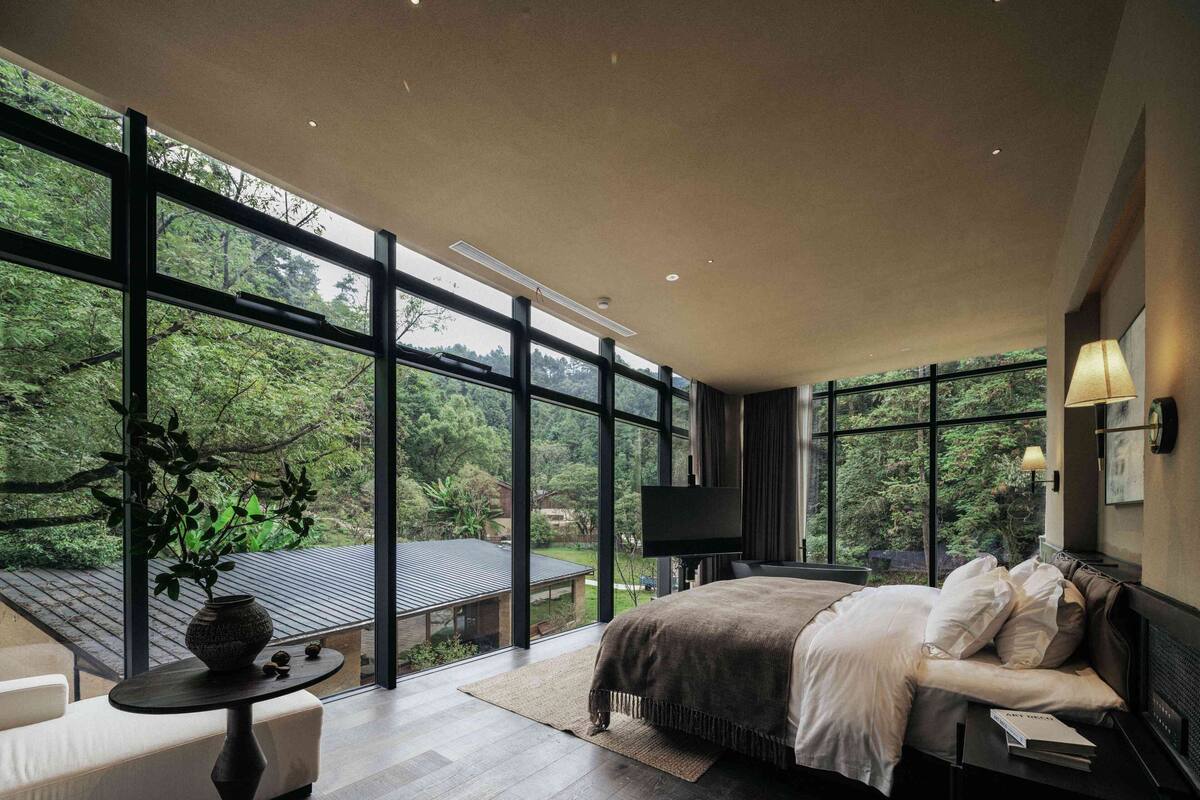 A modern bedroom with a large bed, a sofa, a small table, and a TV. It features large windows offering a view of a garden.