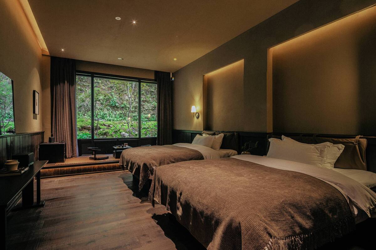 A hotel room with two beds, a large window with a view of greenery, a bedside table, and a lamp.