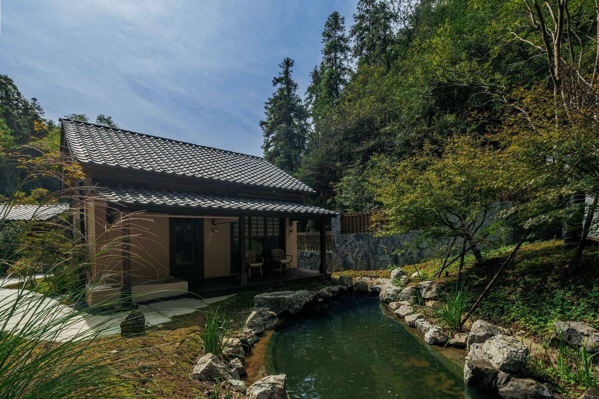 A traditional Japanese-style house with a tiled roof, a koi pond, and a forested backdrop.