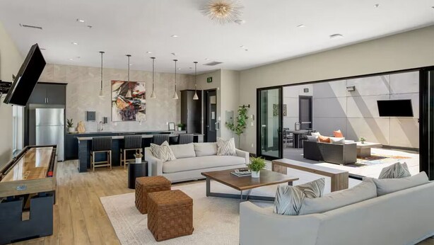 Un salon moderne avec un canapé, une table basse et des poufs. Il y a également un coin cuisine équipé d’un réfrigérateur, d’une cuisinière et d’une table à manger. La pièce est dotée de grandes fenêtres et d’un luminaire au plafond.