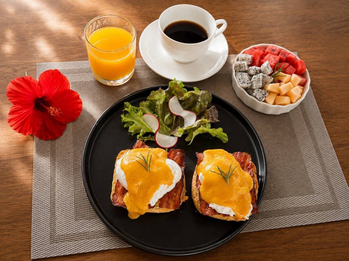 Een ontbijt bord met Eggs Benedict, een glas sinaasappelsap, een kop koffie, een schaal fruit en een rode hibiscusbloem op een tafel.