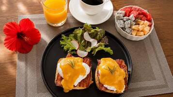 Een ontbijt bord met Eggs Benedict, een glas sinaasappelsap, een kop koffie, een schaal fruit en een rode hibiscusbloem op een tafel.