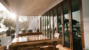 Een houten tafel en stoelen in een zonnige kamer met grote ramen en uitzicht op bomen.