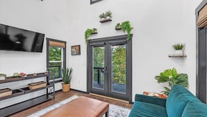 A modern living room with a flat-screen TV, a sofa, and a coffee table. There are potted plants on shelves and a sliding glass door leading to a balcony. - Silent Princess, Tiny Home (Tracy City)