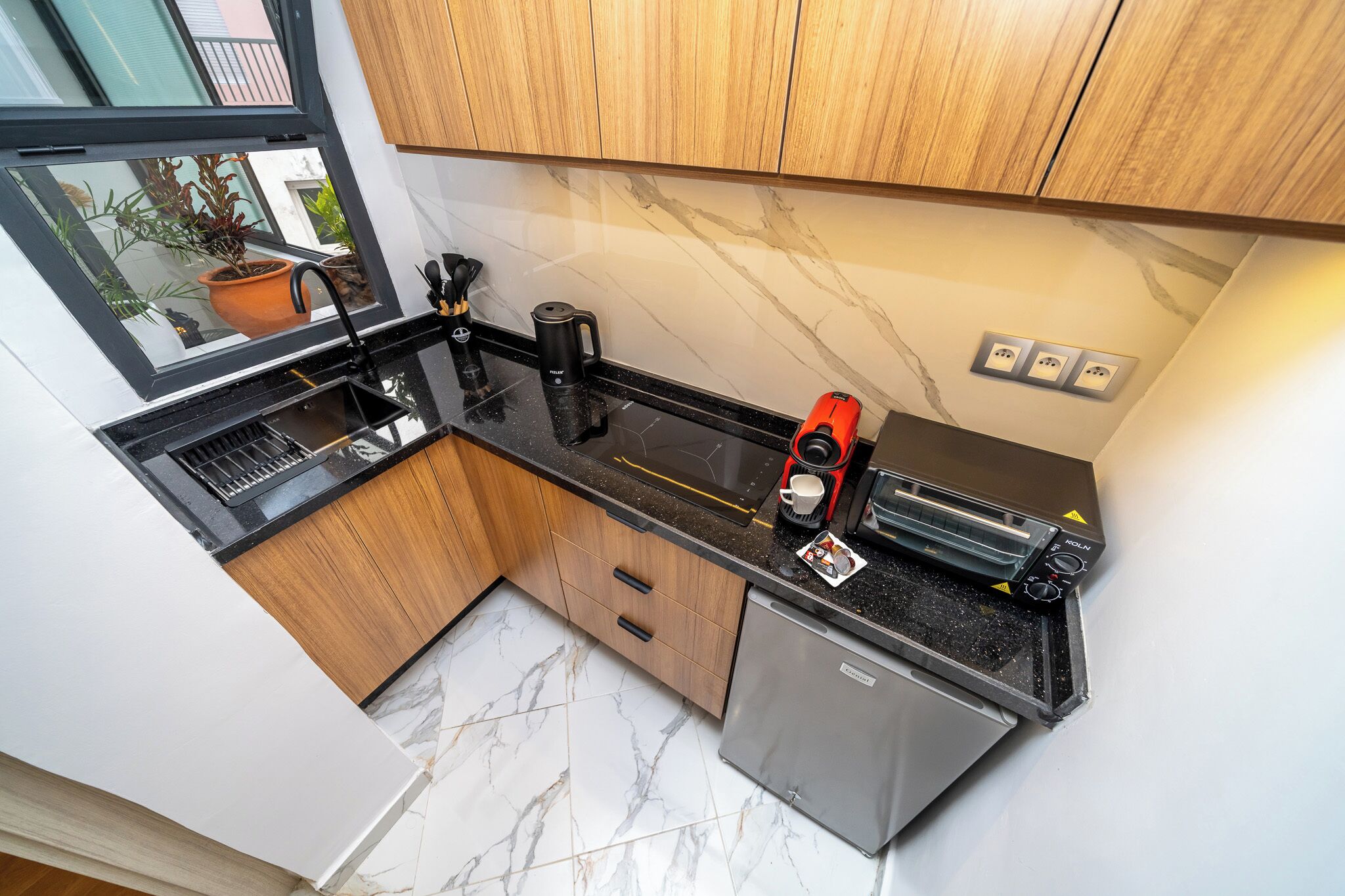 A compact kitchen with wooden cabinets, a black countertop, a coffee maker, a toaster, and a small refrigerator.