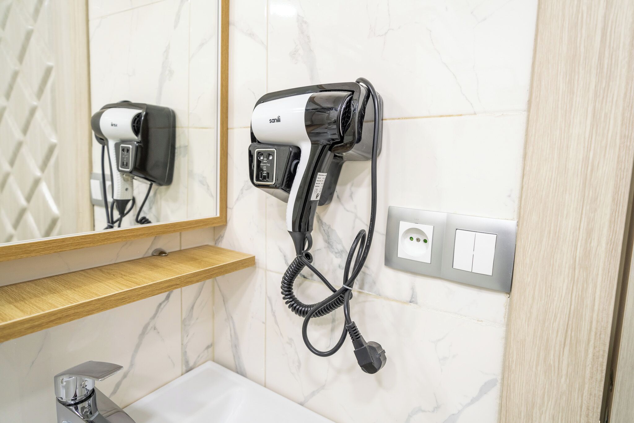 A bathroom with a hairdryer mounted on the wall, a mirror, a wooden shelf, and a sink.