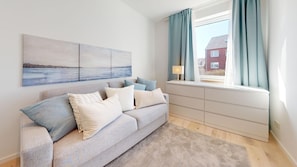 Ein Wohnzimmer mit grauem Sofa, einem beigen Teppich, einem Fenster mit Vorhängen und einem Bild an der Wand.