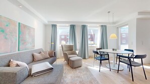 En moderne stue med en grå sofa, et rundt spisebord med stole og et stort vindue med gardiner.
