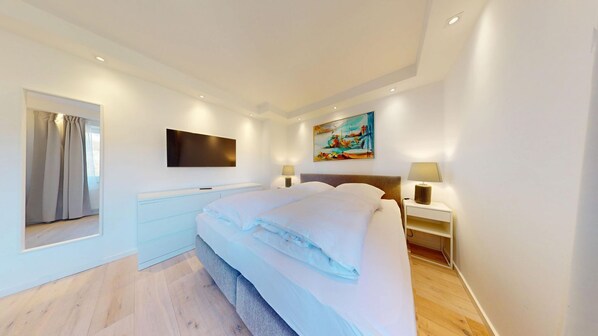 A modern bedroom with a large bed, a flat-screen TV mounted on the wall, and a painting above the bed. - Modern, stylishly renovated 3-room apartment with balcony near the beach - (Hörnum (Sylt))
