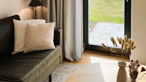 Una sala de estar con un sofá, una lámpara, un florero con flores secas y vistas a un jardín a través de un ventanal corredero.