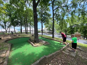 Un campo de mini golf con superficie verde, rodeado de árboles y un borde de piedra.