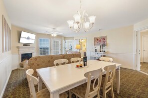 A dining area with a white table and chairs, a sofa, a fireplace, and a television mounted on the wall.