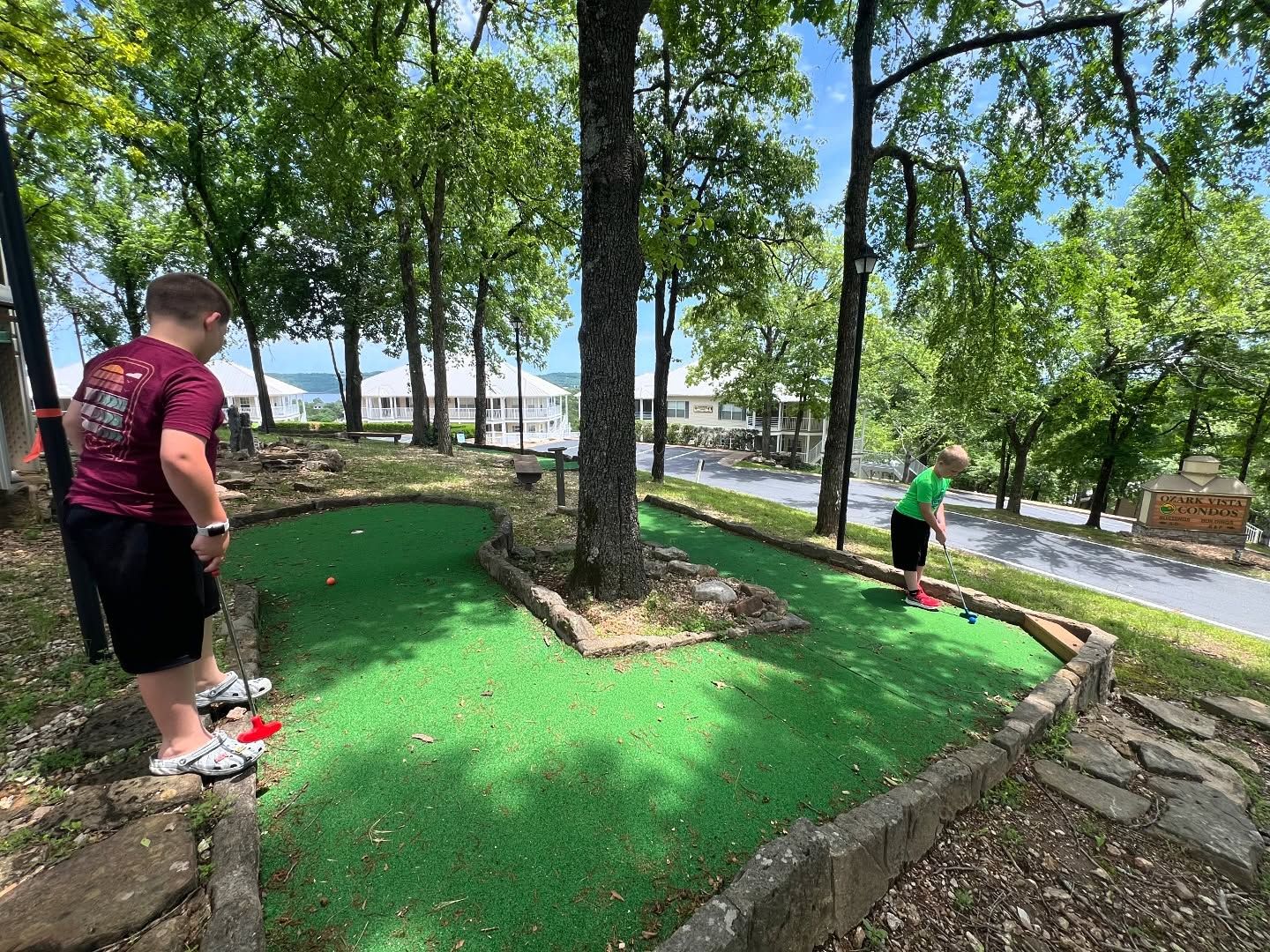 Ein Minigolfplatz mit grüner Spielbahn, umgeben von Bäumen und einem Gebäude im Hintergrund.