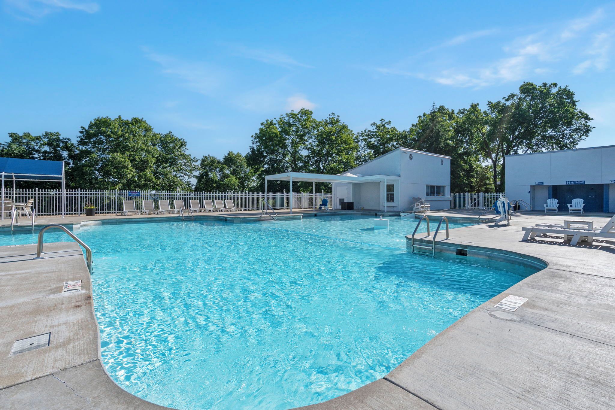 A swimming pool with a white building in the background, a covered seating area, and a fenced area with lounge chairs.