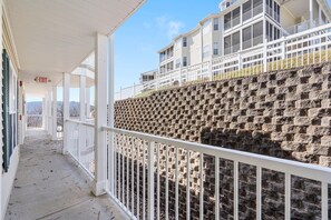 A covered outdoor area with a white railing, a stone wall, and a building with multiple windows and balconies.