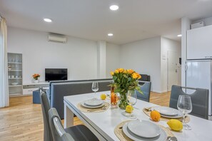 Un coin repas moderne avec une table blanche dressée pour quatre personnes, comprenant des assiettes, des couverts et des verres. Un vase avec des fleurs jaunes est posé sur la table, et on aperçoit un téléviseur au fond.