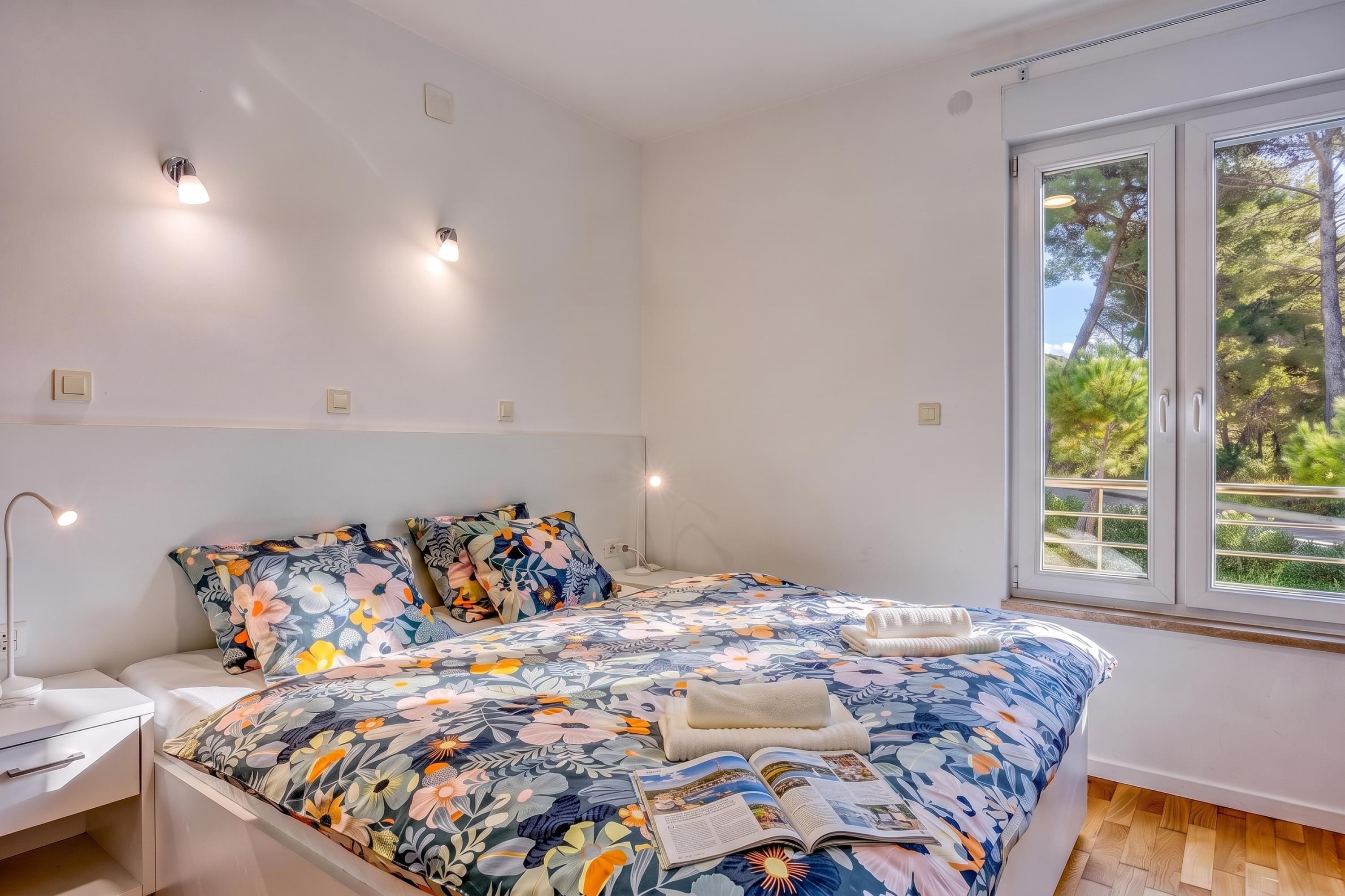 Une chambre à coucher avec un lit, des tables de chevet, une lampe et une fenêtre donnant sur des arbres.