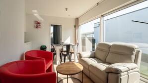 A modern living room with a sofa, red chairs, a small table, and a view through large windows.