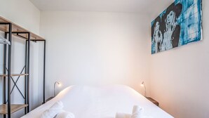 A neatly made bed with white linens and rolled towels, a metal shelving unit, and a painting on the wall.