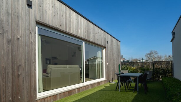 Una casa moderna con amplios ventanales correderos, un patio con mesa y sillas, y un césped cuidado.