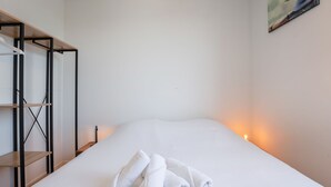 Una cama bien hecha con ropa de cama blanca y toallas enrolladas, una estantería de madera y un cuadro en la pared.