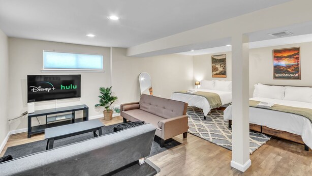 A modern hotel room with a flat-screen TV displaying Disney+ and Hulu, a sofa, a bed, and a bedside table. - 4BR Family Getaway w/ King Bed + 5 TVs, Mins to DC/OldTown,Gameroom,Pet Friendly (Alexandria)
