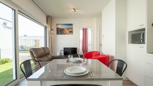 Een moderne eetruimte met een glazen tafel, zwarte en rode stoelen, een televisie en een schilderij aan de muur.
