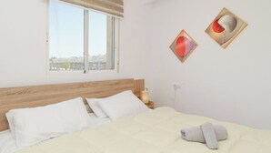 Un dormitorio con cabecera de madera, un ventanal con persianas, dos obras de arte colgadas en la pared y una cama bien hecha con una toalla doblada.