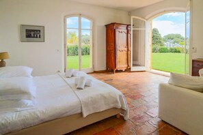 Une chambre avec un grand lit, une armoire en bois et une vue sur un jardin verdoyant, visible à travers une porte ouverte.
