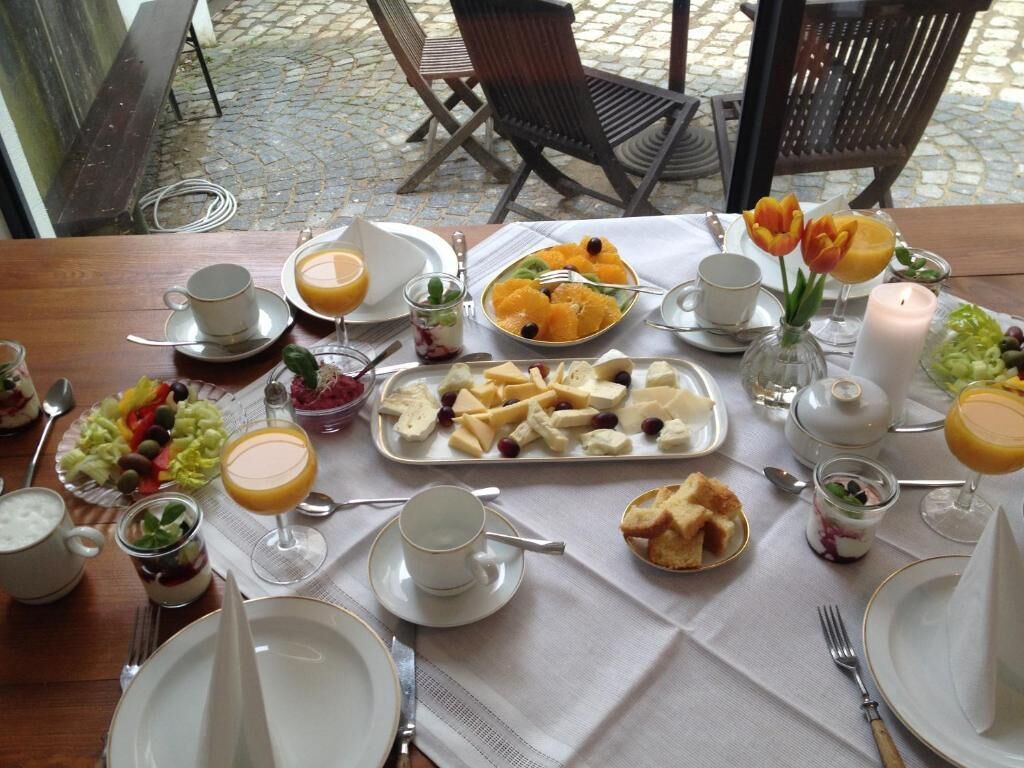 Una mesa de desayuno servida con platos, vasos y una variedad de alimentos, entre ellos frutas, quesos y pan.